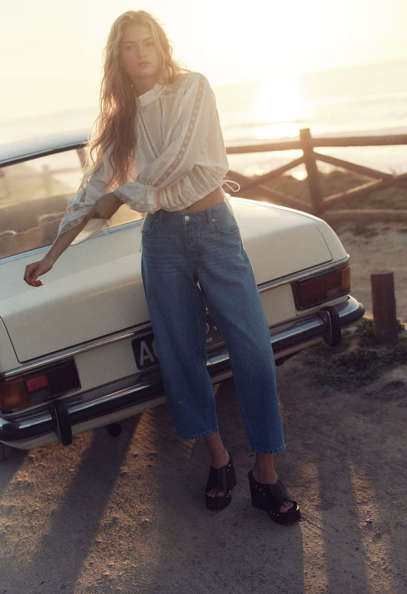 A young woman in a white top and jeans leans against a vintage car with the sunset in the background in a fashion editorial shot.