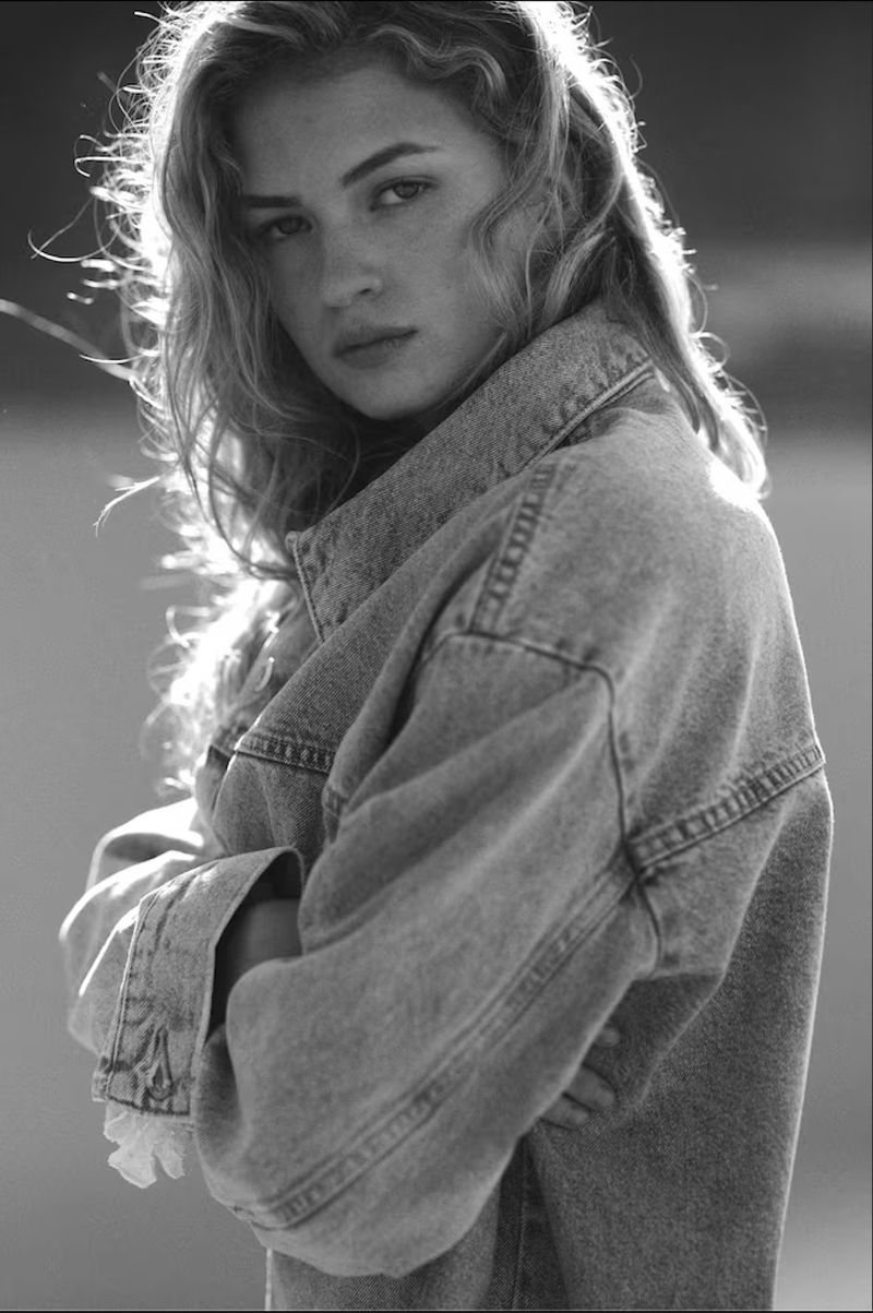 Monochrome portrait of a fair-skinned young woman with curly hair, wearing a denim jacket, standing outdoors with arms crossed.