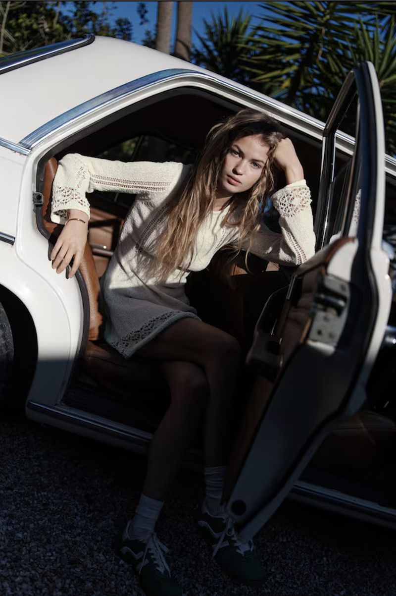 A young woman with long blonde hair in a short white dress leans against a vintage white car with its door open in an outdoor setting.