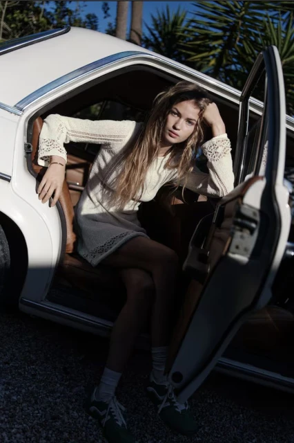 A young woman with long blonde hair in a short white dress leans against a vintage white car with its door open in an outdoor setting.