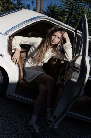 A young woman with long blonde hair in a short white dress leans against a vintage white car with its door open in an outdoor setting.