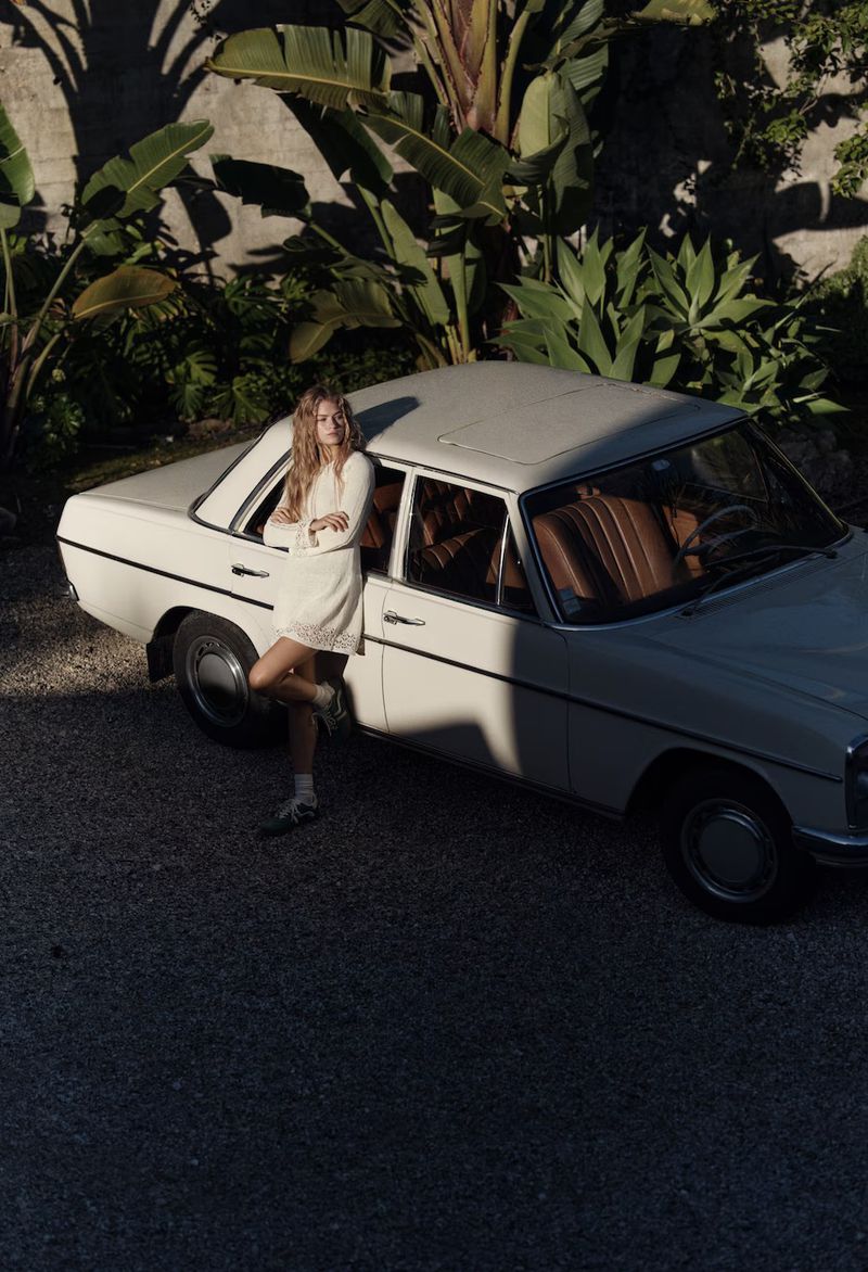 Young woman in a white dress leaning against a vintage white car surrounded by tropical plants in an outdoor setting.