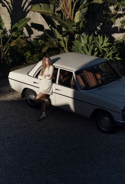 Young woman in a white dress leaning against a vintage white car surrounded by tropical plants in an outdoor setting.