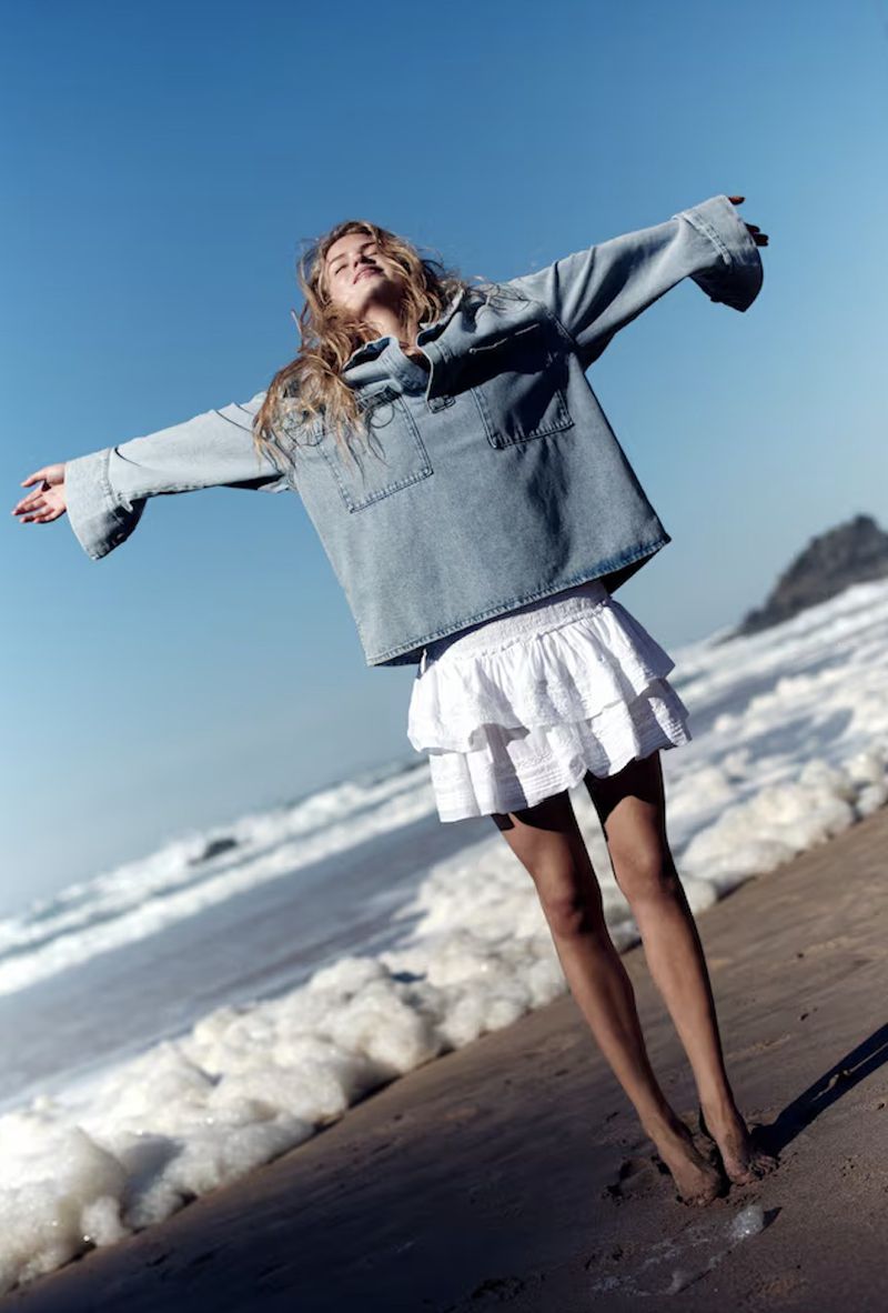 Woman in denim shirt and white skirt stands barefoot on sandy beach with arms outstretched, set against ocean waves and blue sky.