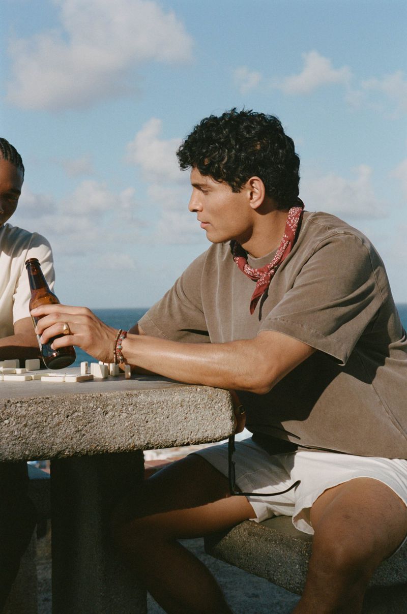 Two men play a board game on an outdoor concrete table near the ocean on a sunny day. One man holds a beer bottle.