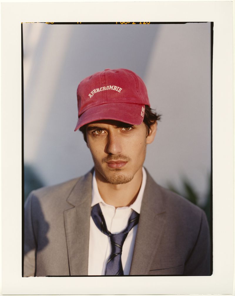 Portrait of a man wearing a red baseball cap, grey blazer, white shirt, and tie against a plain wall.