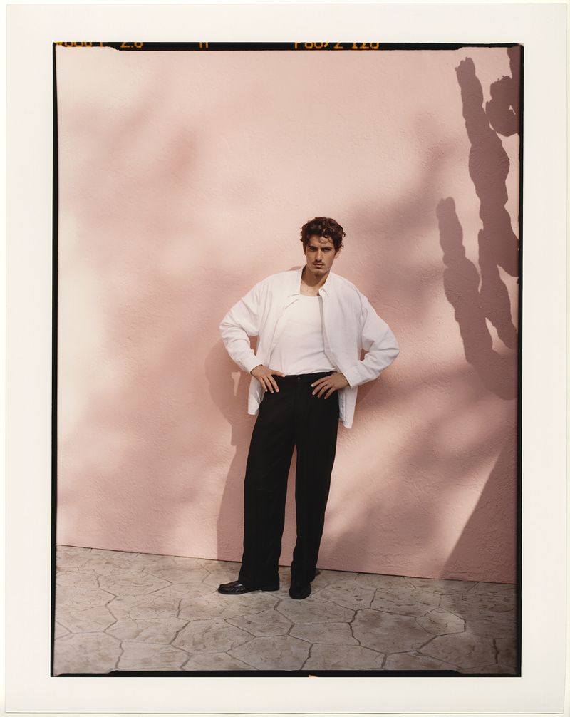 Adult male in white jacket and black pants poses with hands on hips against a pink wall with cactus shadow in a medium shot.