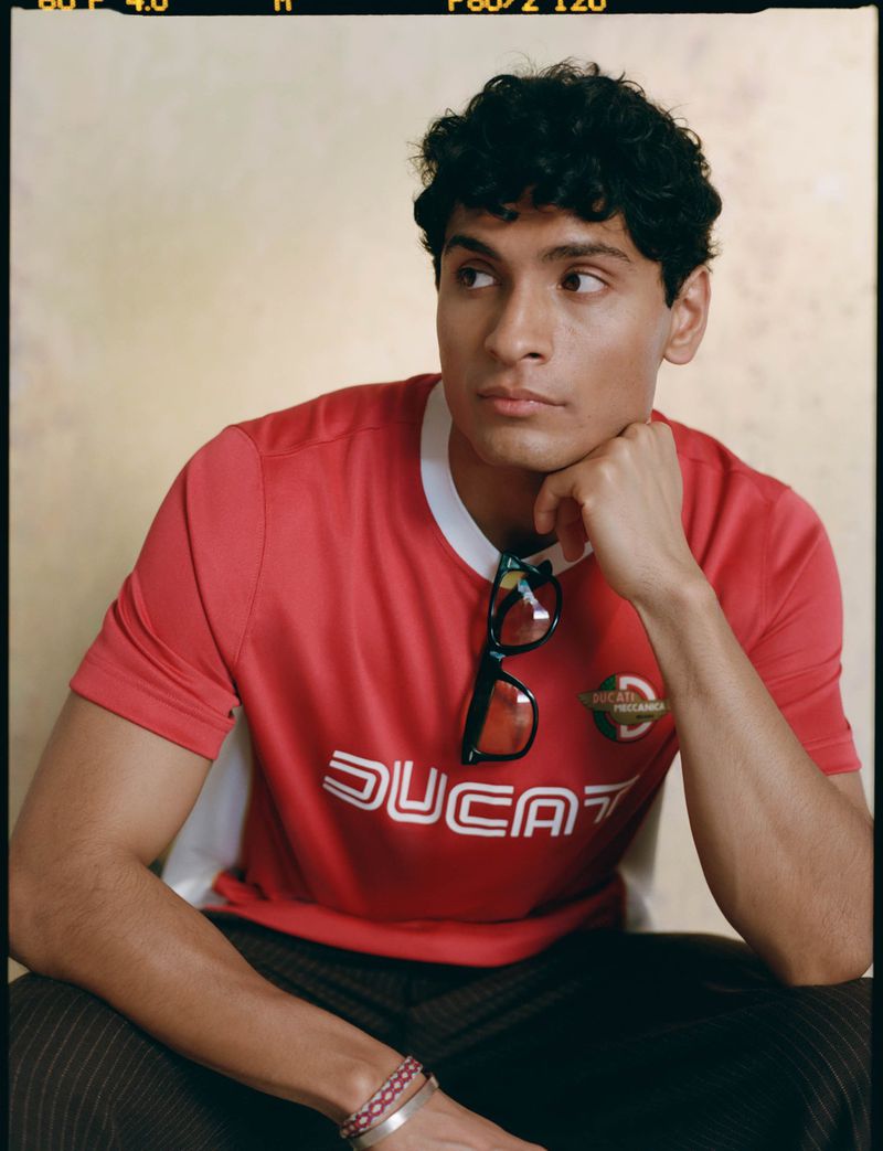 Attractive young man with curly dark hair in red Ducati shirt sitting indoors with glasses. Fashion photography in studio setting.
