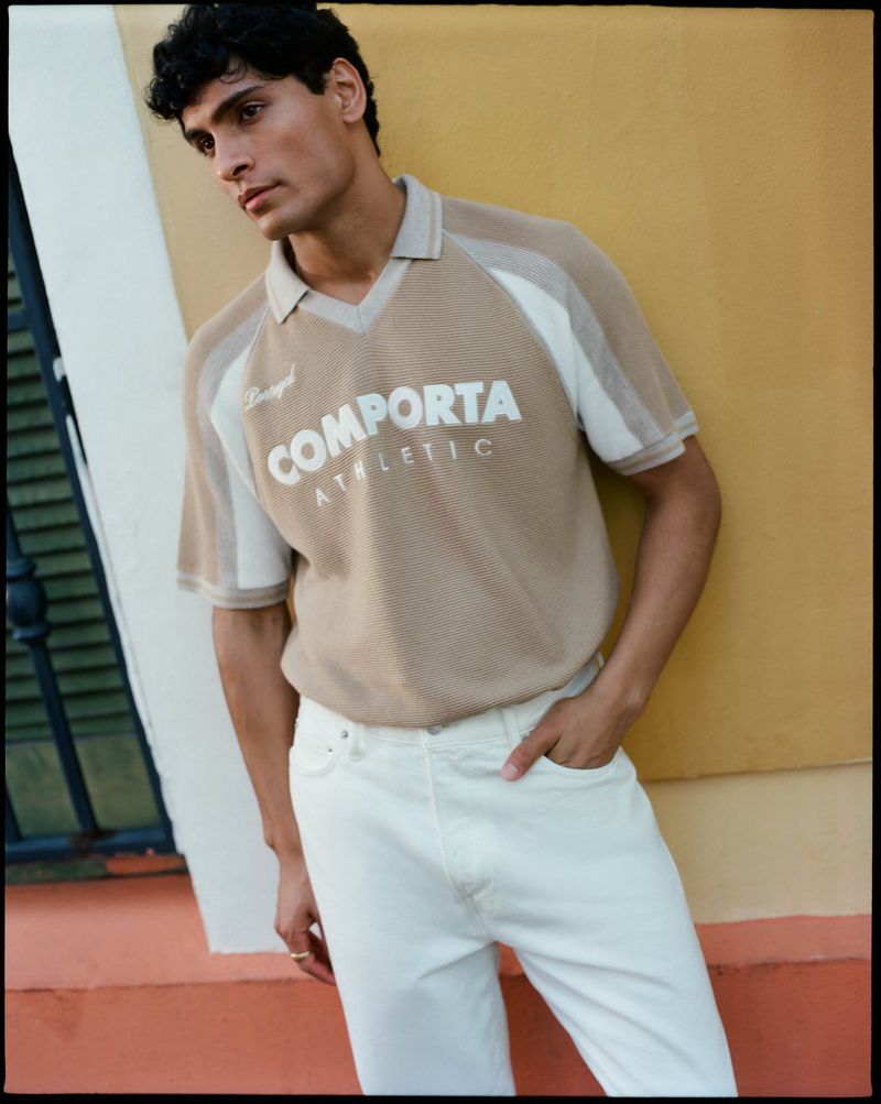 A light-skinned male model poses in white pants and a beige and white collared polo shirt in front of a two-tone wall.