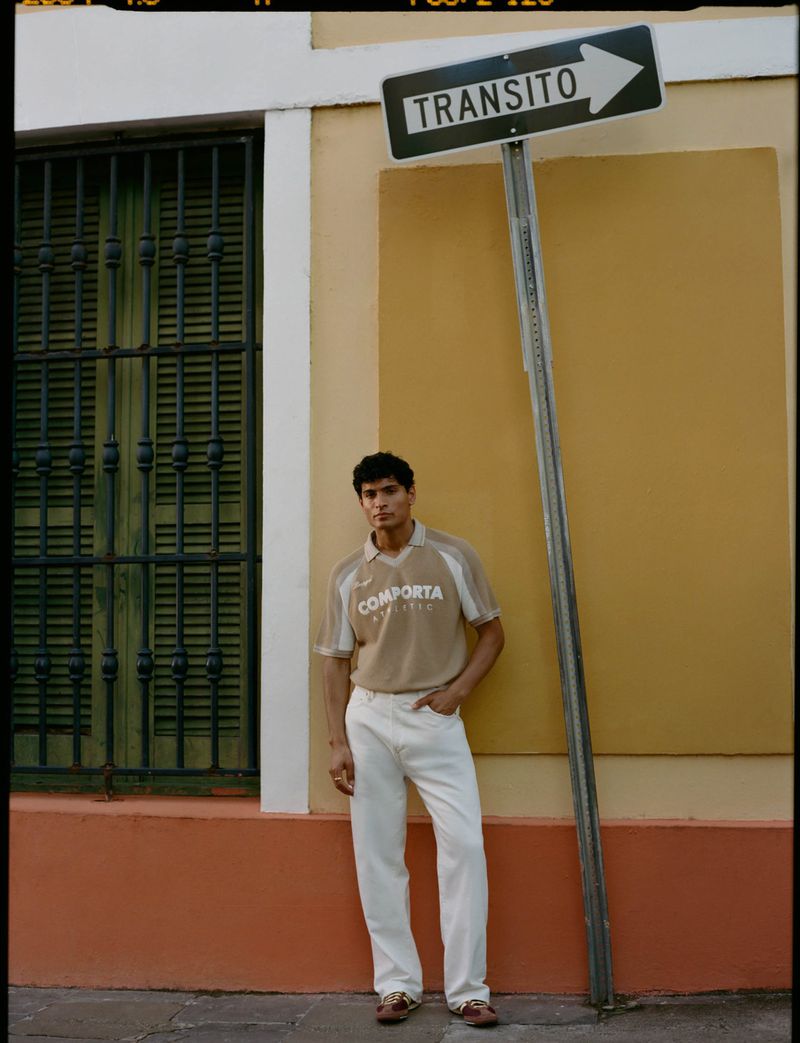 Young adult male stands against a yellow wall next to a 'Transito' sign, wearing a tan shirt and white jeans, shot in natural light.