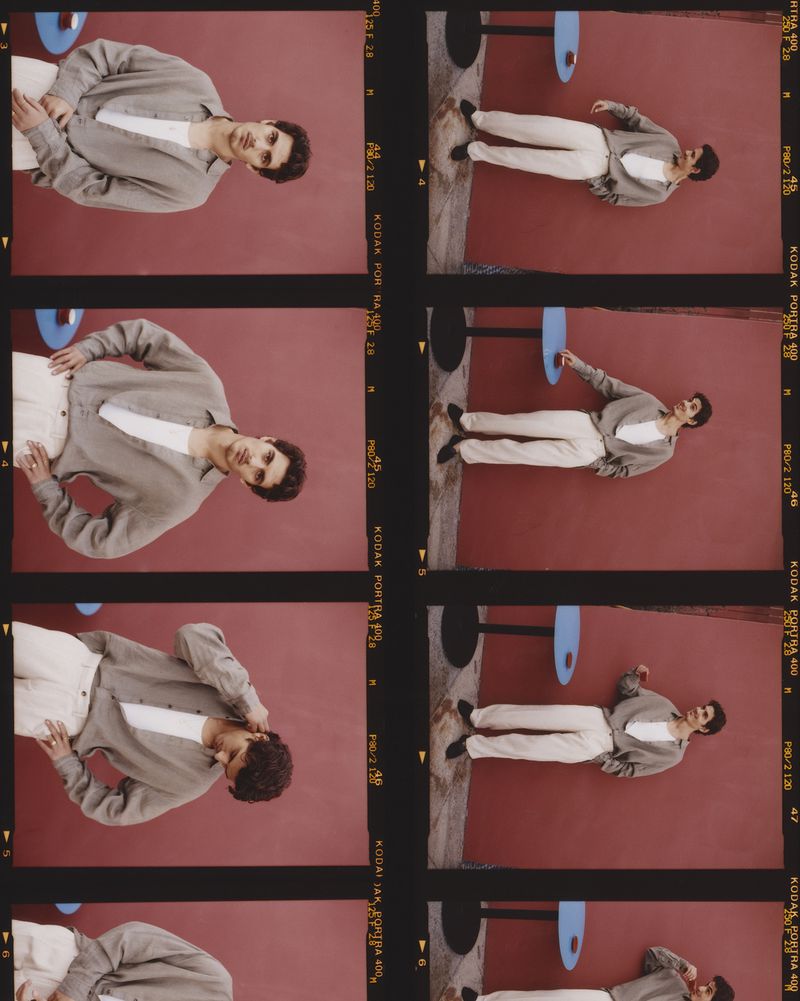 Man poses indoors wearing casual shirt and pants, leaning against a solid wall, captured in a film strip format.
