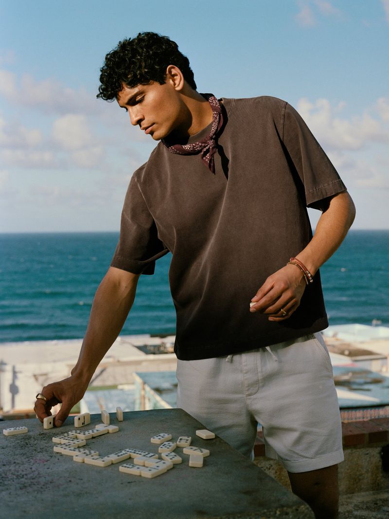 A young adult male plays dominoes outdoors on a rooftop with a bandana around his neck, wearing shorts and a t-shirt; ocean backdrop.
