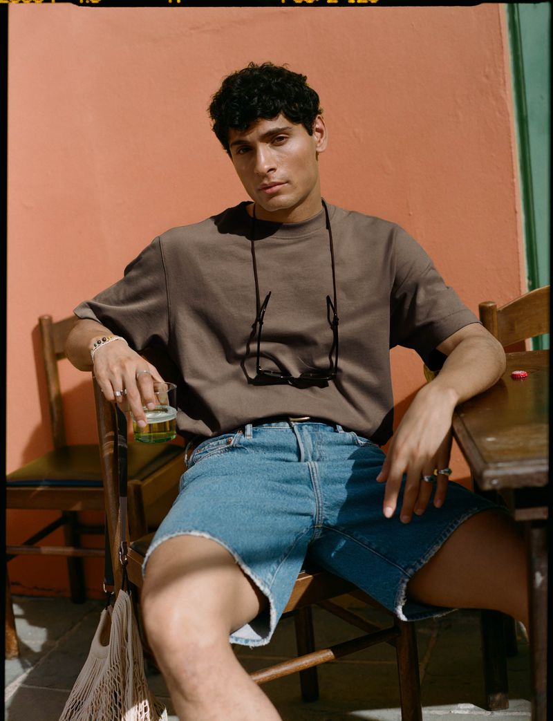 Young adult male poses in denim shorts and a brown t-shirt, sitting at a table outside with peach and turquoise walls.
