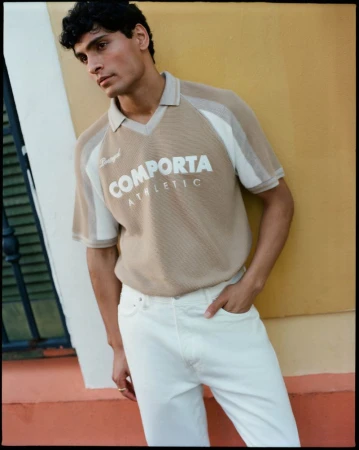 Male model wearing a beige Comporta Athletic polo shirt and white jeans standing against a warm yellow wall.