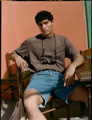 Man in brown t-shirt and denim shorts sitting outdoors holding a glass with a peach-colored wall behind him.