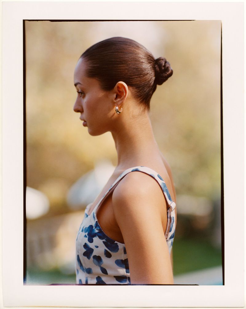 Profile portrait of a young woman with her hair in a bun, wearing a blue and white floral dress with a blurred outdoor background.