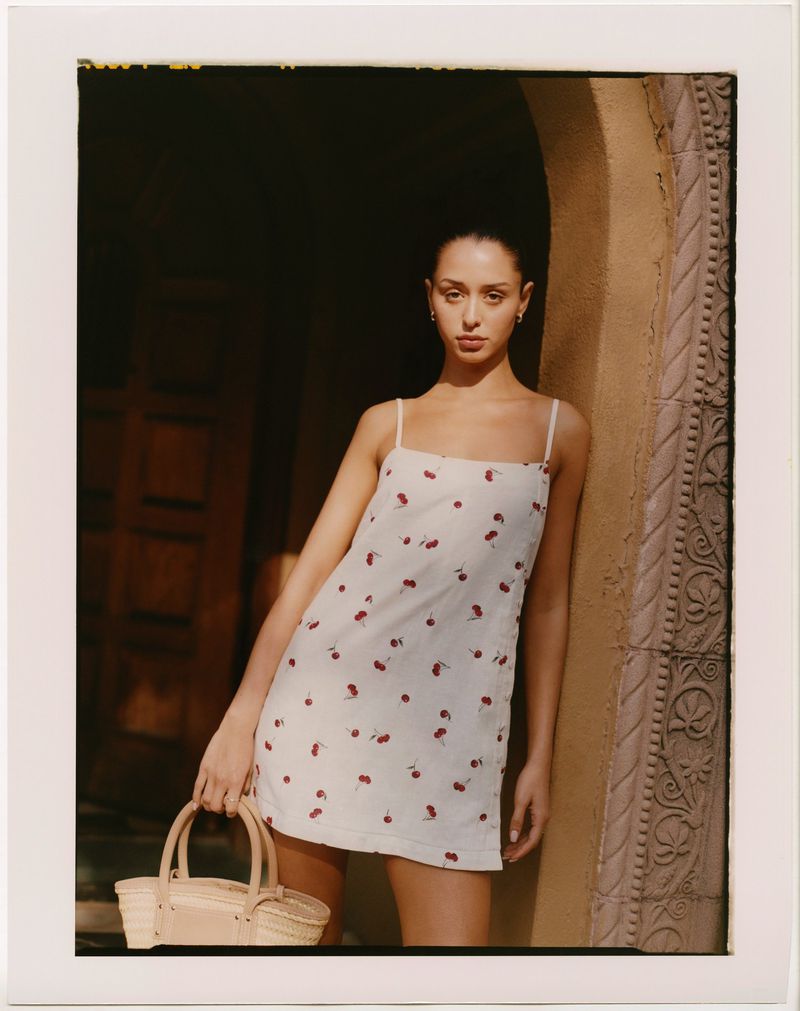 Woman in white cherry-print dress holds a straw basket bag in a tan archway with ornate carvings. Natural light and neutral tones.