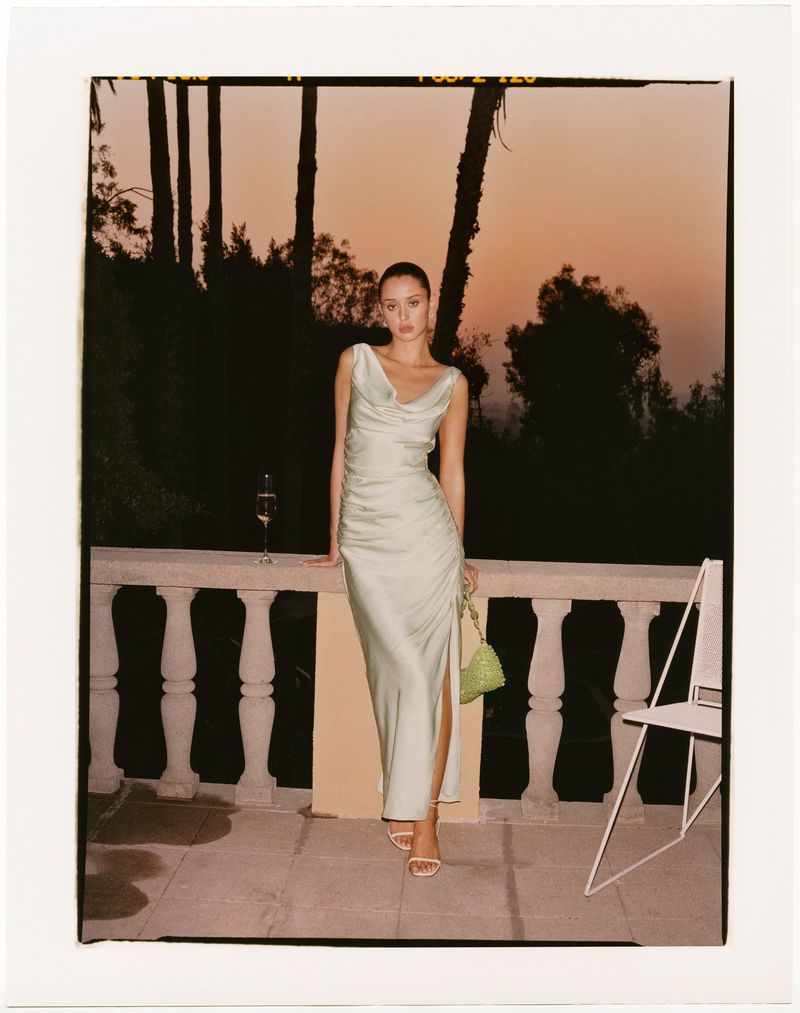 A woman in an evening gown stands on a terrace at sunset, holding a clutch, beside a drink and a white chair. 
