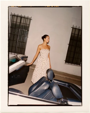 Elegant woman in polka-dot dress poses with a classic white convertible car in an outdoor setting against a wall with barred windows.