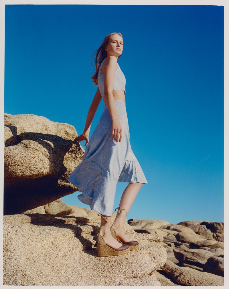 A young adult woman poses in a blue two-piece outfit on a rocky beach under a clear blue sky, showcasing fashion and outdoor style.