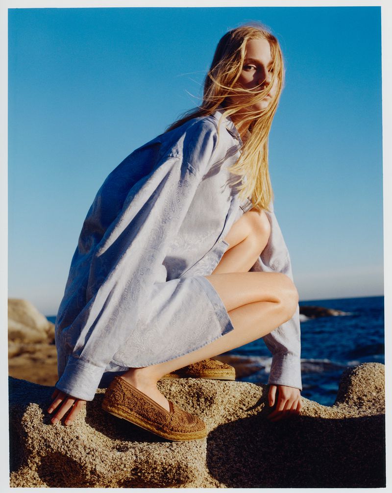 Fashion shot of a young adult female model in a blue shirt and brown shoes, squatting on rocks near the sea under a clear blue sky.
