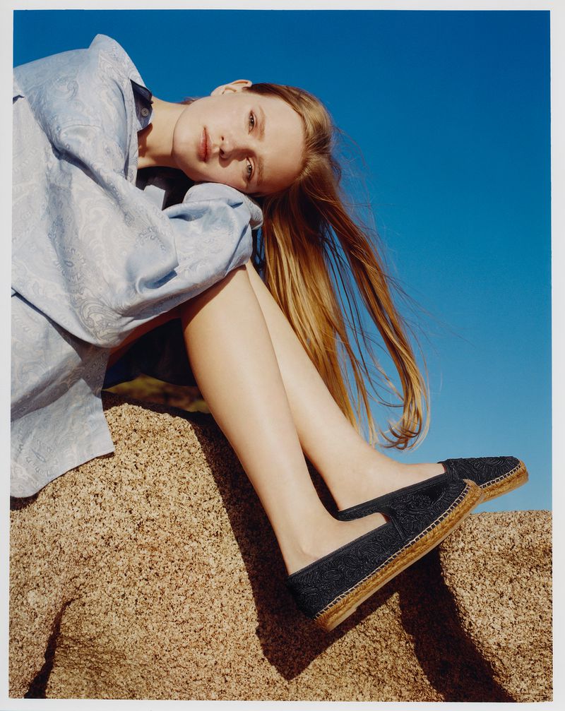 Young woman with long brown hair resting on a rock wearing slip-on shoes and a blue patterned shirt under a clear blue sky.