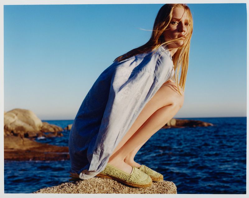 Woman poses on a rock at the beach wearing a light blue shirt dress and green espadrilles, ocean and sky are in the background.