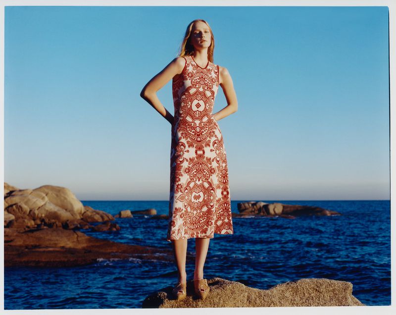 A fair-skinned woman poses on a rocky shore in a sleeveless, patterned maxi dress, with the blue sea and sky in the background.