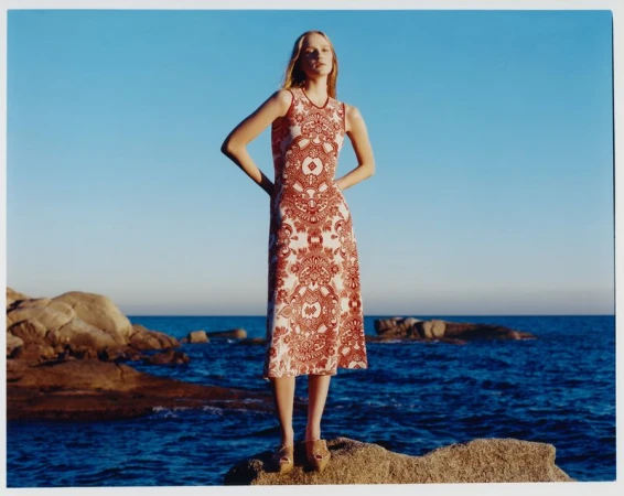 A fair-skinned woman poses on a rocky shore in a sleeveless, patterned maxi dress, with the blue sea and sky in the background.