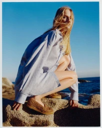 Model crouching on coastal rocks wearing espadrilles and an oversized shirt against an ocean view