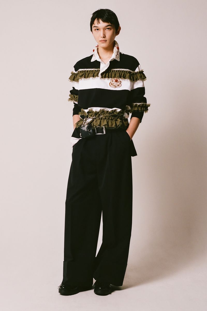 Full-length studio shot of a person with dark hair wearing a black and white striped ruffle-trimmed shirt and wide-leg black pants.