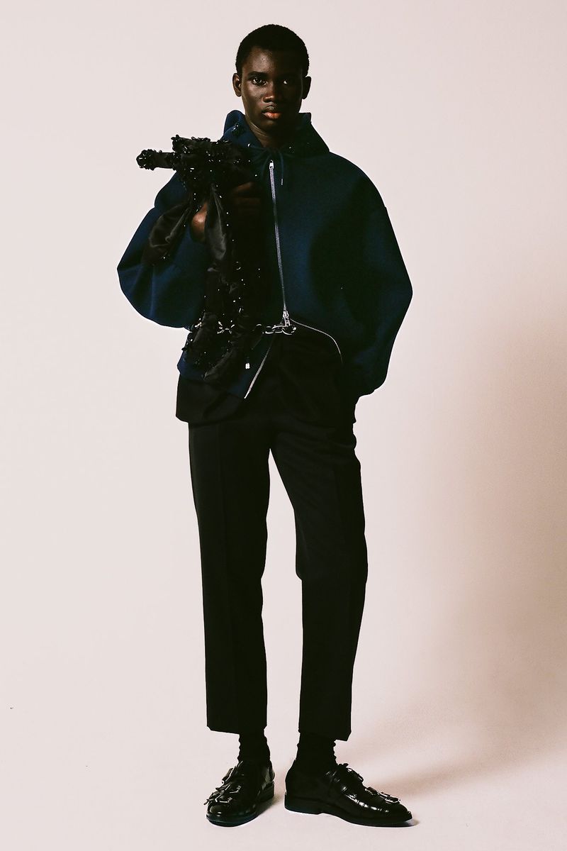 A stylish person in a navy hooded zip-up jacket and black pants poses confidently in a studio, holding a textured decorative piece.