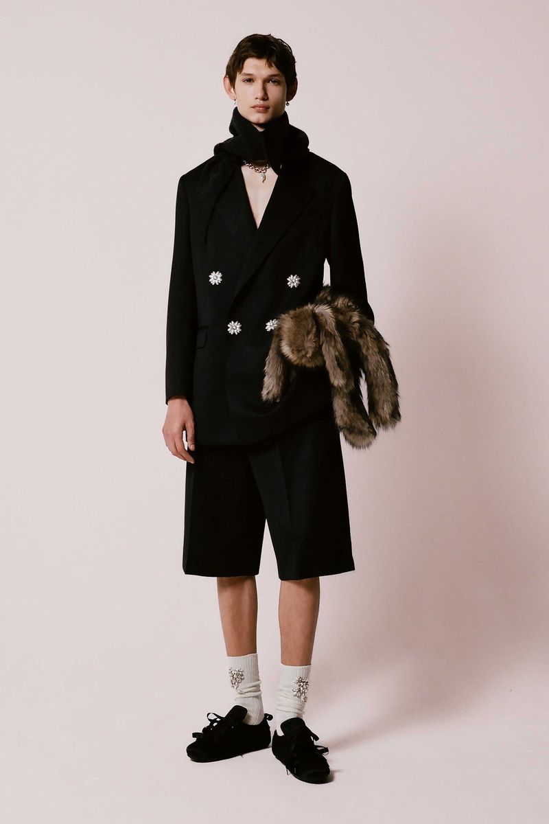 A fair-skinned male fashion model poses in a black suit, shorts, and scarf with black shoes in a studio setting for a fashion editorial.