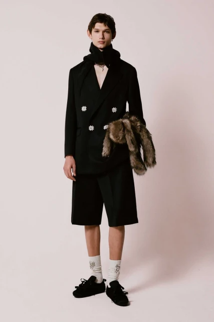 A fair-skinned male fashion model poses in a black suit, shorts, and scarf with black shoes in a studio setting for a fashion editorial.