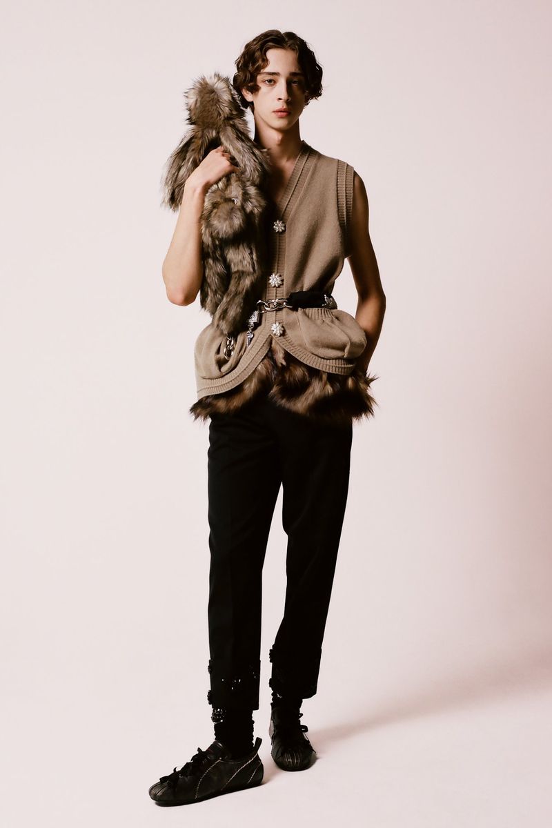 Full-length studio portrait of a young man in a sleeveless tan top and black pants, carrying a fur pelt over his shoulder.