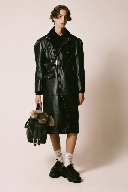 A slender man in a long black leather coat, black shoes, and socks, poses with a fur-trimmed backpack against a plain backdrop.