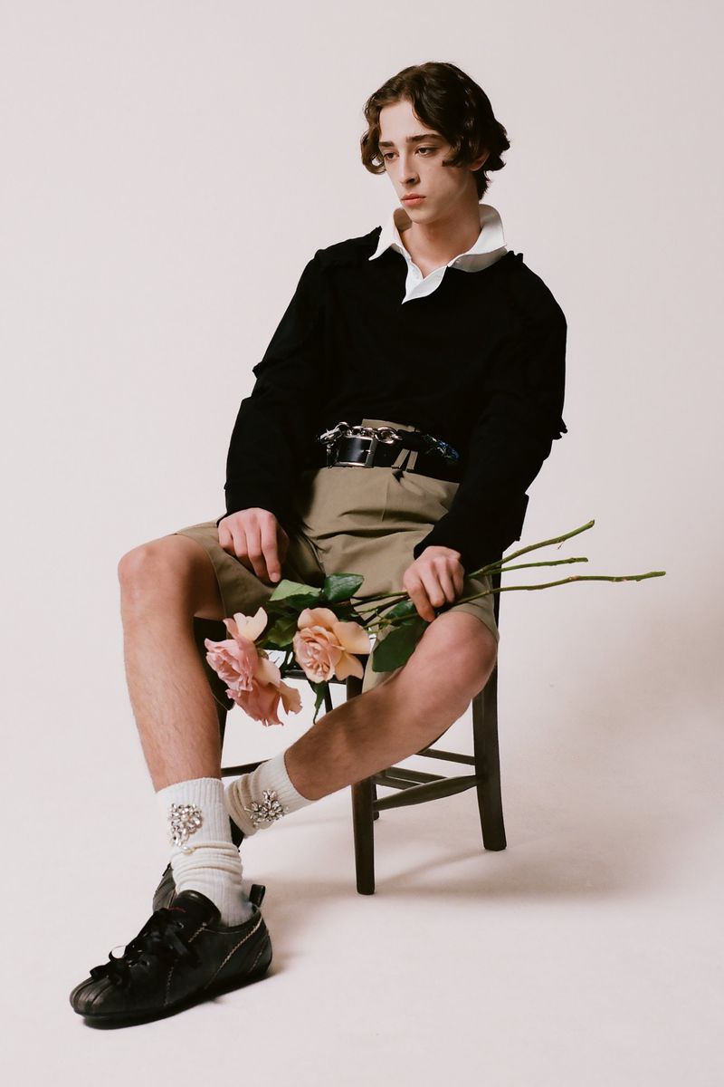 A young man sitting on a wooden chair holding pale pink roses against a plain background for Simone Rocha F/W 25