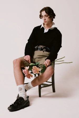 A young man sitting on a wooden chair holding pale pink roses against a plain background for Simone Rocha F/W 25