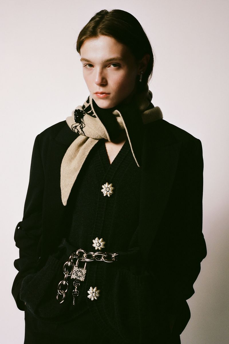 Portrait of a young adult posing in a black cardigan with floral buttons and a beige scarf with a chain around the waist.