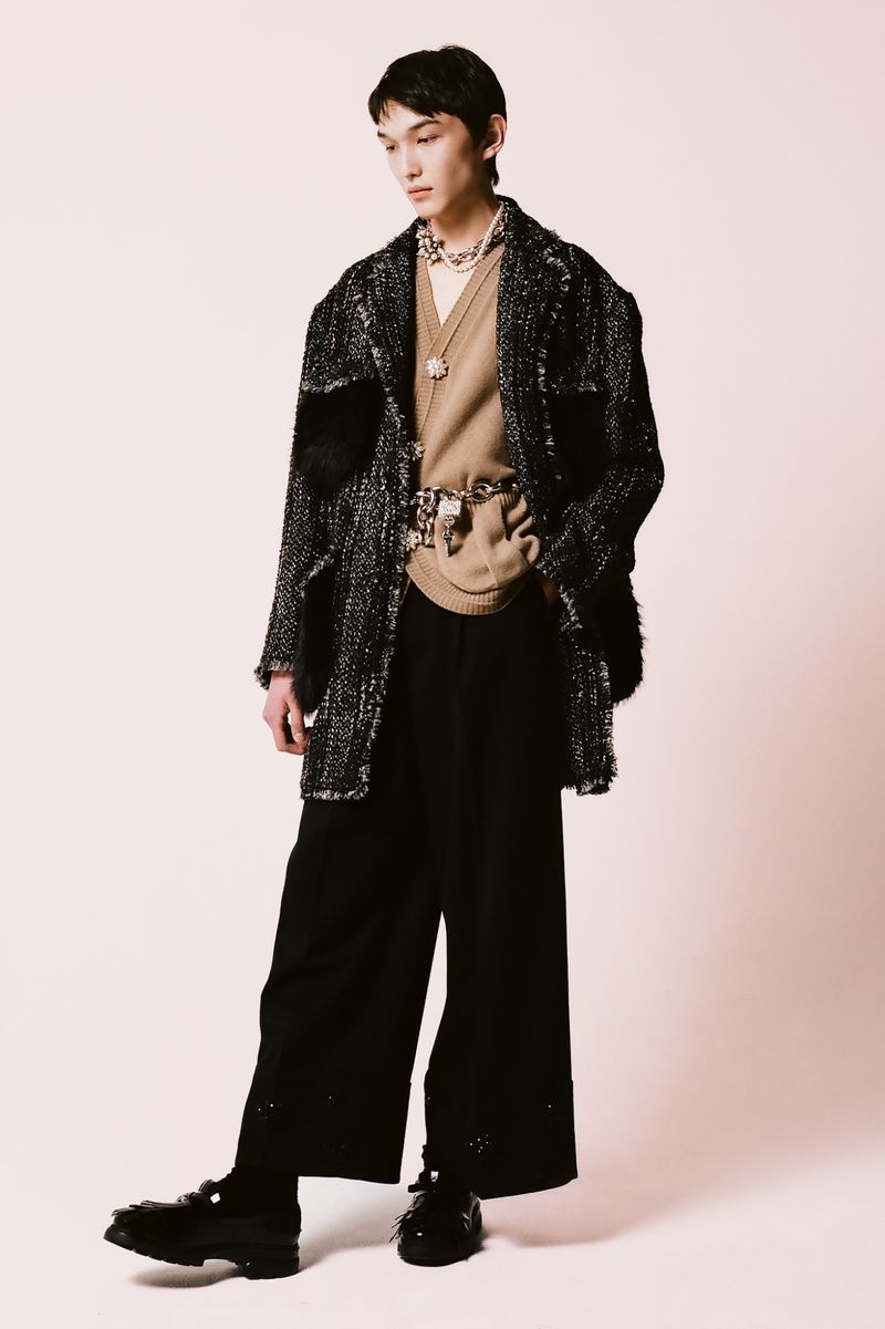 Asian male model in a beige sweater and dark wide-leg pants wearing a dark textured overcoat poses against a light backdrop.