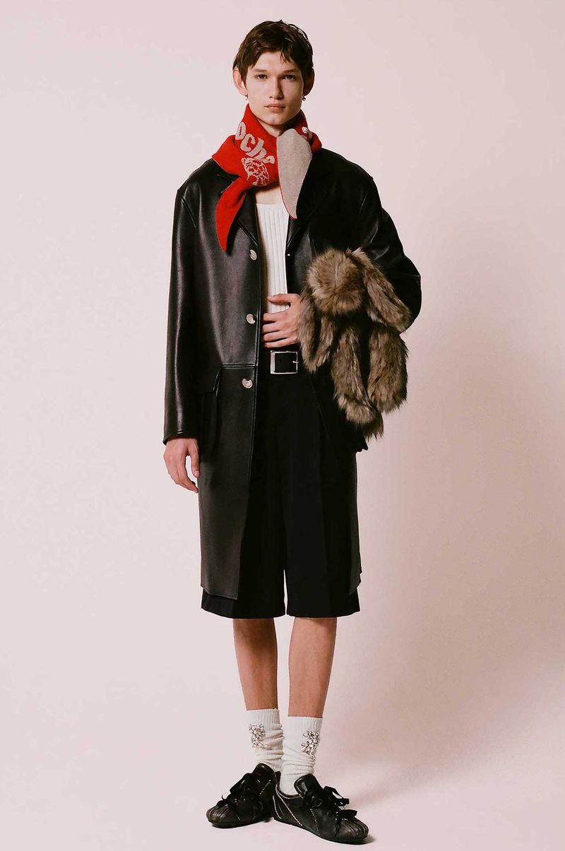 Fashionable adult male poses indoors wearing black coat, shorts, socks and shoes; accessorized with red scarf and plush toy.