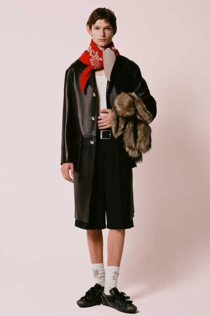 Fashionable adult male poses indoors wearing black coat, shorts, socks and shoes; accessorized with red scarf and plush toy.