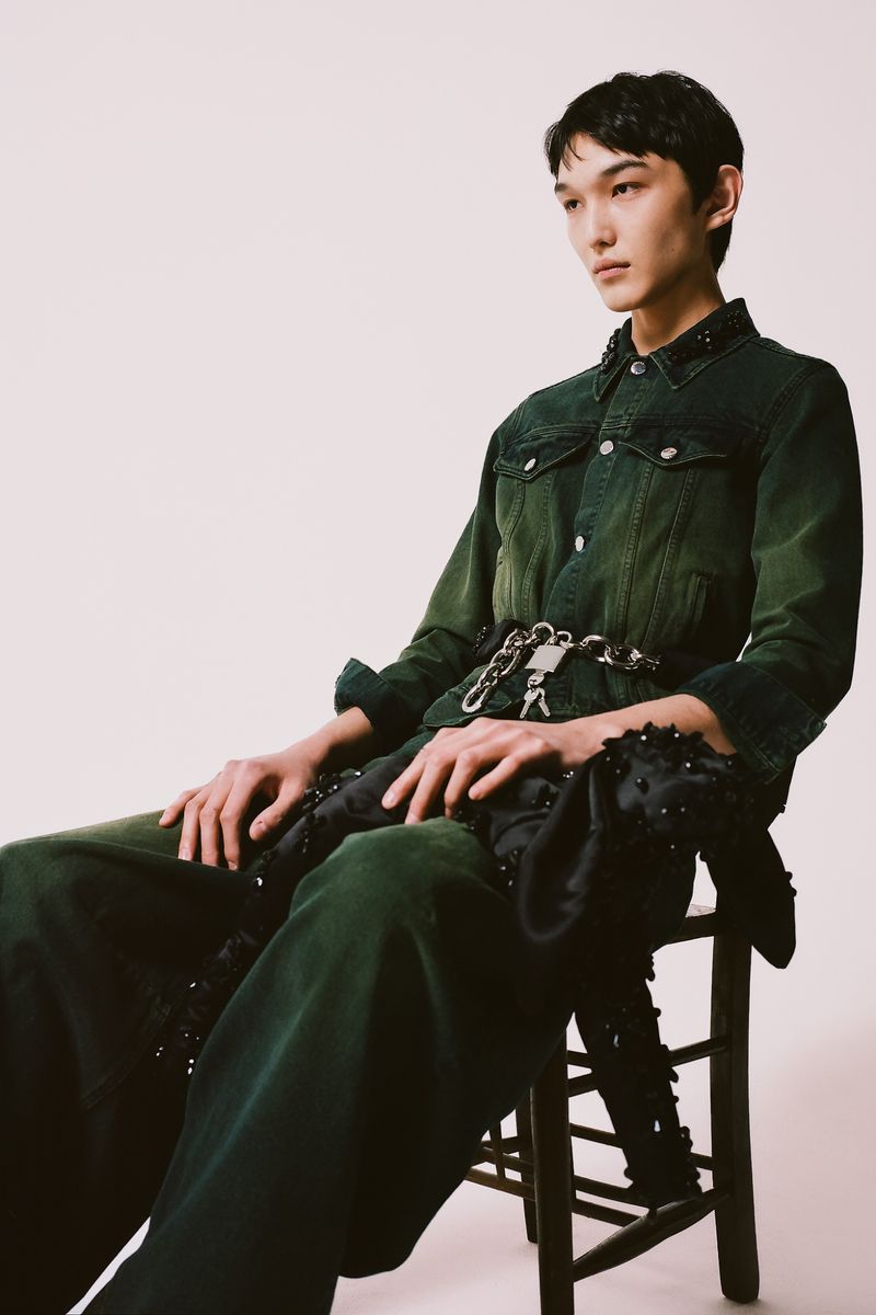 Young man in a dark green denim outfit sits on a wood stool in a studio setting, accessorized with a metal chain and padlock belt.