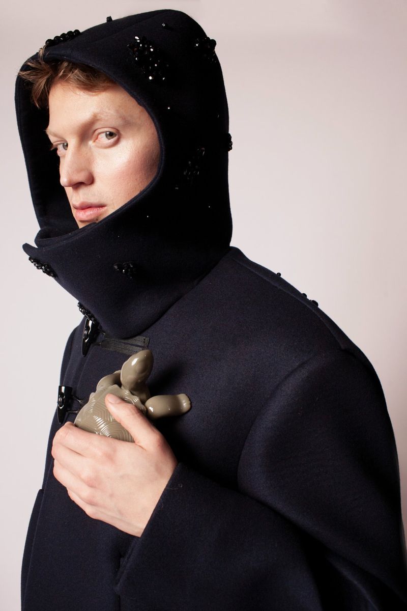 Fashion portrait of a person wearing a navy hooded coat with embellishments, holding a pale grey prosthetic hand indoors against a white backdrop.
