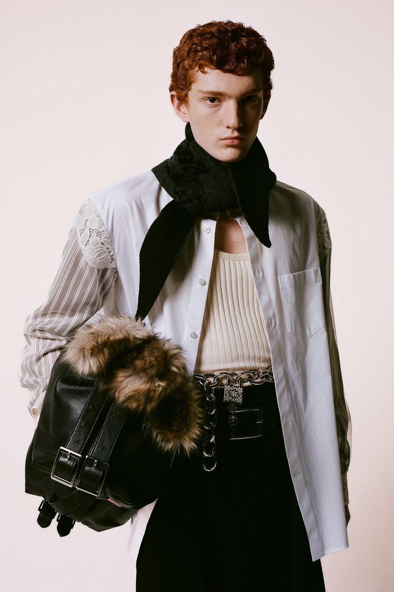 Androgynous fashion model with red hair wears a black scarf, white shirt, and carries a fur-trimmed bag in a studio portrait.