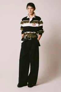 Model wearing a black and white striped polo shirt with ruffle detailing and black trousers against a neutral background