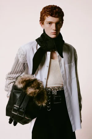 Model with red hair wearing a white button-down shirt, dark scarf, and carrying a black bag with fur trim against a plain background