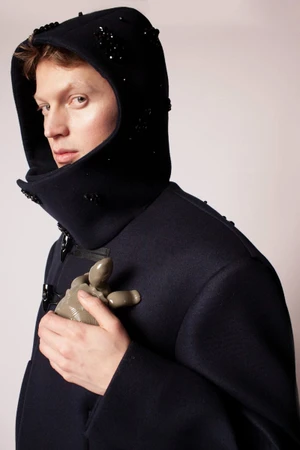 Model wearing a dark hooded duffle coat with bead embellishments, holding a ceramic object against a seamless background.