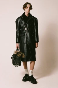 Male model wearing a long black leather coat holding a dark handbag against a plain white seamless studio background.