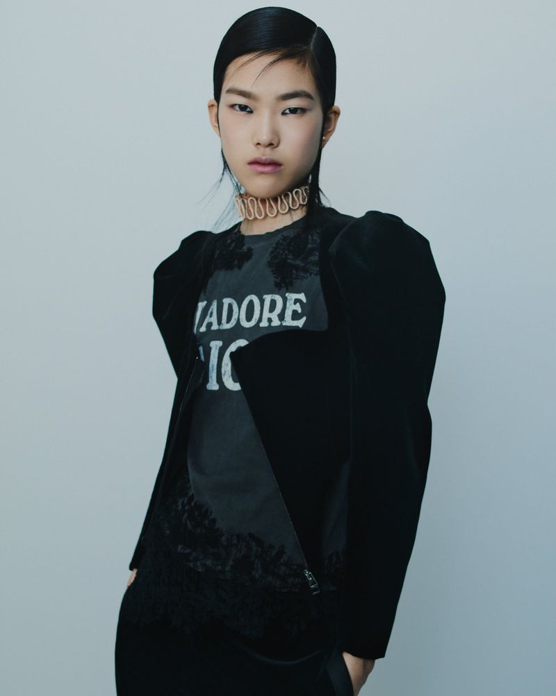 Asian woman in a black velvet jacket and t-shirt with text graphic against a light grey background in a studio fashion shot.